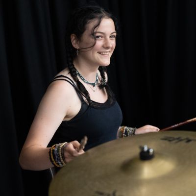 A girl with brown hair sits behind a red drum kit and smiles